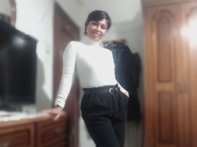 JessiHt with short black hair, white ribbed turtleneck, and black high-waisted pants, standing with hands in pockets, smiling, blurred image.