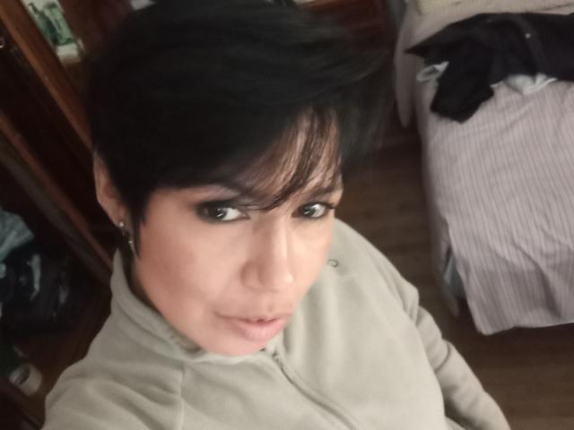 JessiHt with short black hair, brown eyes, light skin, wearing a beige hoodie, looking up at the camera with a slight smile, in a dimly lit room.