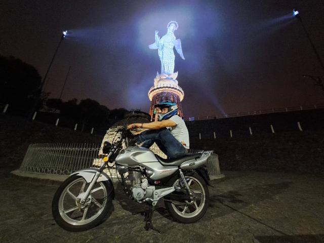 Davydwolf-hot on a silver motorcycle, wearing a helmet, with a statue of a blue-lit goddess on a tall pedestal illuminated from above in the night.