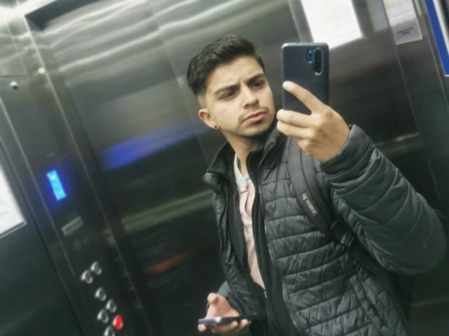 Muso69 taking a selfie in an elevator, wearing a black quilted jacket, looking annoyed, holding a phone and a card, with short black hair, and small ear studs.