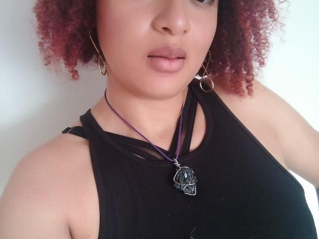 VioletaSwitch, com cabelos ruivos e cacheados, vestindo uma blusa preta sem mangas, um colar roxo com um pingente preto e brincos de argola dourados, tem os lábios levemente entreabertos..