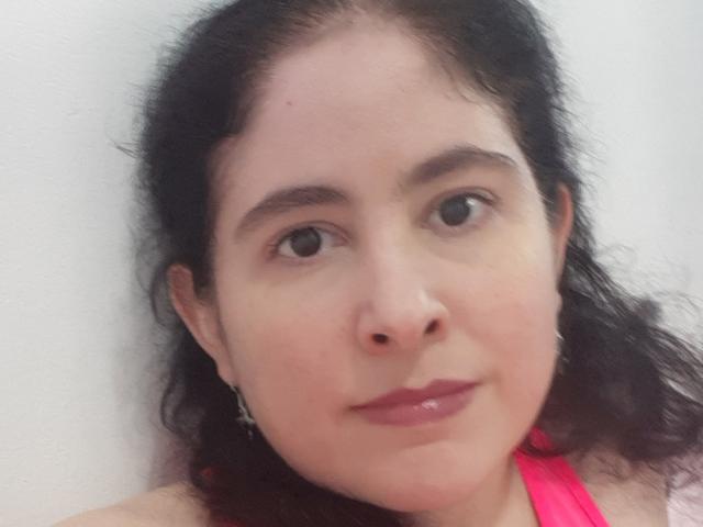 PamyToLike has dark curly hair, brown eyes, pink lipstick, and a light skin tone. She's wearing a pink top with a cross earring on her left ear.