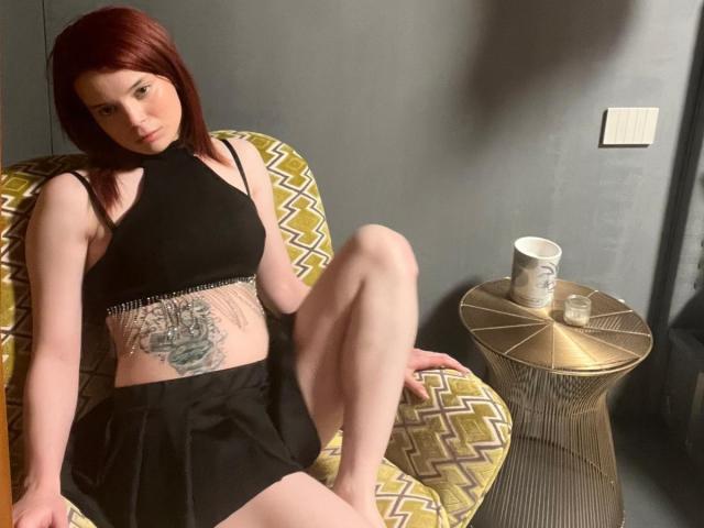 Yumeko69 with red hair, black crop top, and black shorts, sitting on a patterned chair, showing a tattoo on her midriff, with her legs up and a gold table nearby.
