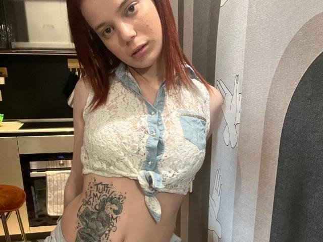 Yumeko69 with red hair, wearing a white lace sleeveless top, tied at the front, exposing a tattoo on her left side and her small, perky tits. She's in a kitchen.