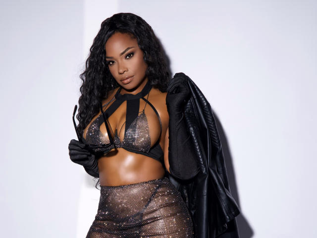 RachelDiamond with dark skin, curly black hair, wearing a sheer black mesh top exposing her big tits, black high-waisted mesh skirt, black gloves, and black leather jacket.