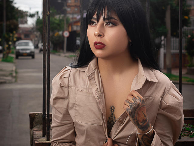 NebulosaI with black hair, tan shirt, red lipstick, tattooed hand, and cleavage, sitting outdoors, looking up with a serious expression, urban street background.