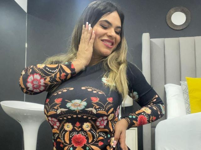 BrianaPreston with large tits, wearing a black floral dress, smiling, hand on cheek, blonde ombre hair, black eyebrows, red lipstick, white nails, gray headboard, yellow pillow.