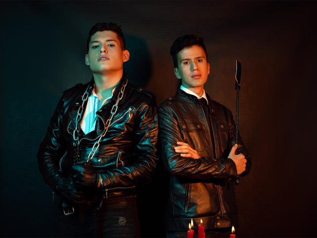 Two DomStravelers in black leather jackets stand back-to-back, one with a chain, the other holding a whip, both with arms crossed, candles in front, dark, moody lighting.