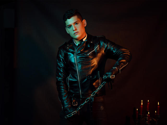 DomStravelers with short dark hair, wearing a shiny black leather jacket and holding a chain, standing in dim light with red candles in the background.