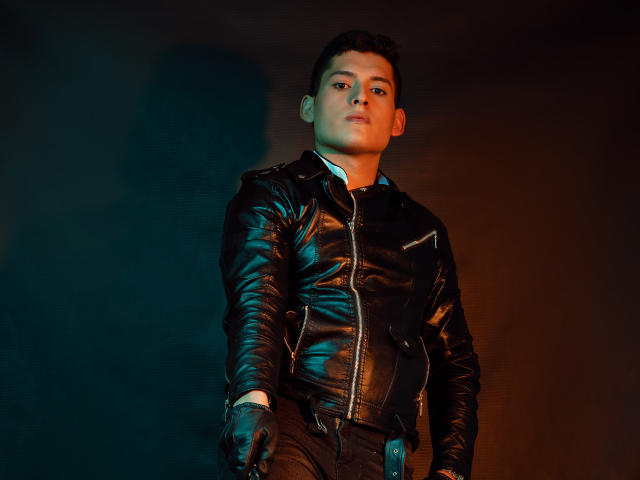 DomStravelers with short dark hair, wearing a shiny black leather jacket and gloves, standing against a dark background, looking confidently to the side with a slight smirk.
