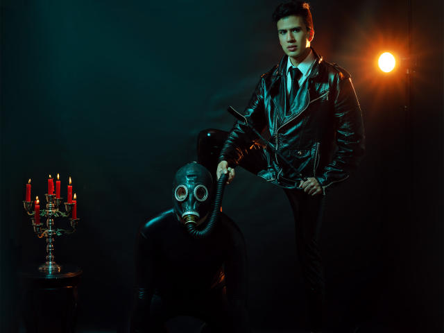 DomStravelers in black leather jacket and white shirt, holding a gas mask over another DomStraveler's face, wearing a similar mask, in front of a candelabrum with red candles.