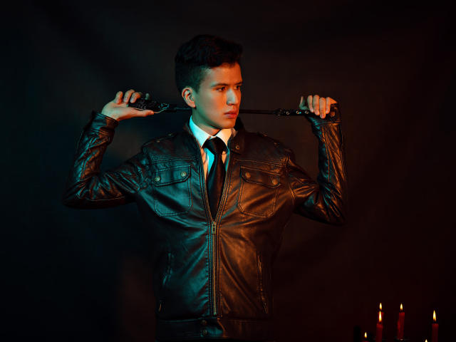 DomStravelers with dark leather jacket, holding a whip over his shoulder, wearing a black tie, white shirt, and greenish-black light. Red candles in the bottom right.