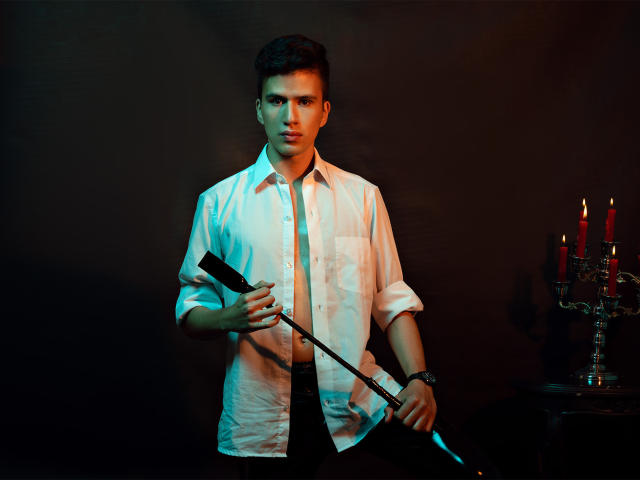 DomStravelers with short dark hair, wearing a white button-up shirt, holding a black whip and a black tablet, standing with a dark background and a candelabrum on the right.