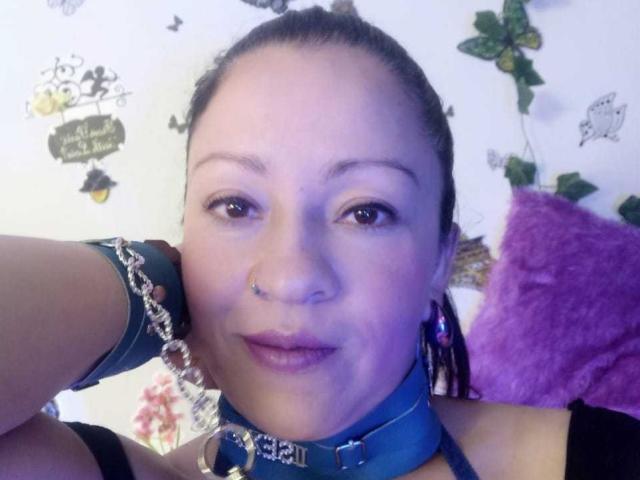 CharloteEzra with a nose ring, blue leather collar with metal chain, and matching wrist cuffs, brown eyes, and purple lipstick, looking at the camera.