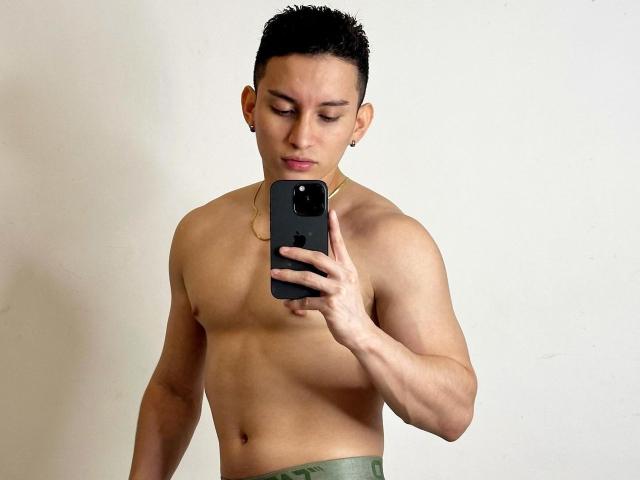 SantiagoXoxo, shirtless, holding a phone, taking a mirror selfie, showing a muscular, toned upper body, short black hair, and a gold necklace, wearing green pants.