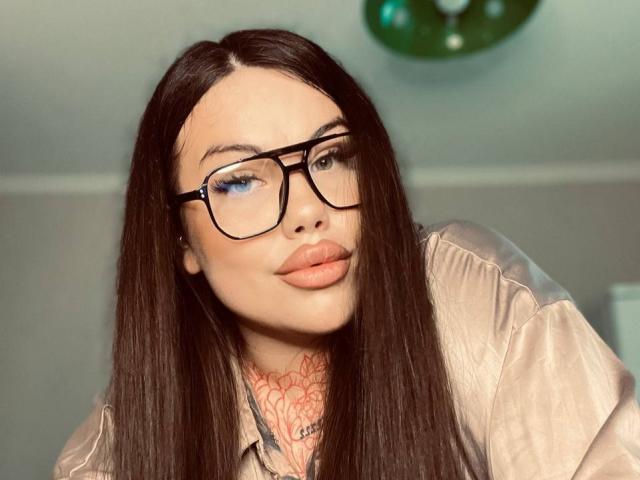 SpicyPeach has long brown hair, black glasses, blue eyeshadow, and is wearing a shiny beige shirt. Her tattooed neck and plump lips are visible.