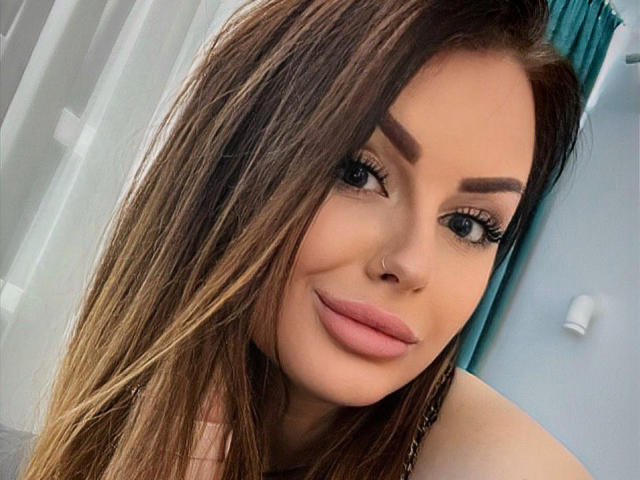 MiaHotOh has long brown hair, thick eyebrows, long eyelashes, pink lips, and a small nose ring. She's smiling with a cute, sexy look.