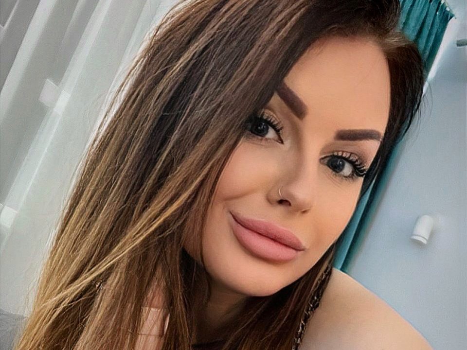 MiaHotOh has long brown hair, thick eyebrows, long eyelashes, pink lips, and a small nose ring. She's smiling with a cute, sexy look.