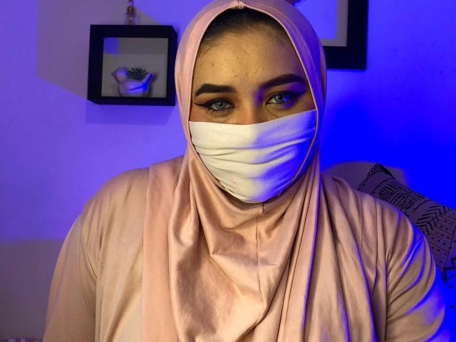 AishaGhulam with a pink hijab, white mask, and matching pink top, has blue-lit eyes and thick eyebrows. She's looking directly at the camera, showing a confident expression.