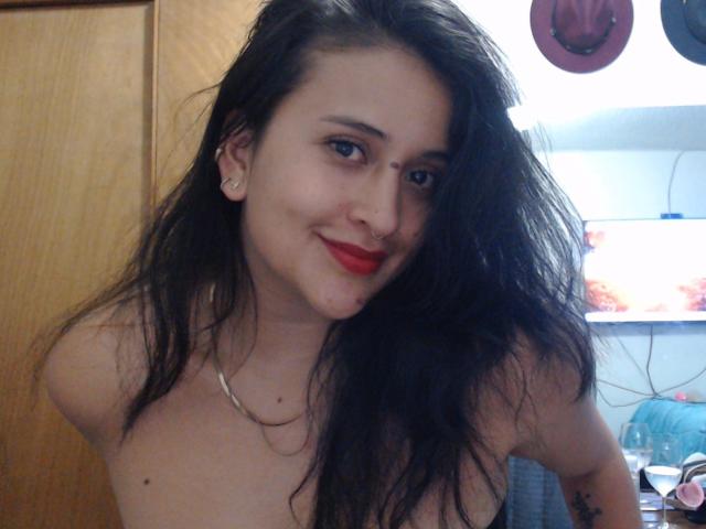 GiovannaBellaC with wet black hair, red lipstick, nose ring, and earrings, smiling, topless, showing small tits, and a tattoo on her right arm, in a dim room.