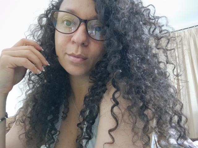 Angelonix, avec ses cheveux noirs bouclés, ses lunettes et un léger sourire, dévoilant ses petites seins dans un haut à décolleté bas, posant sa main sur sa joue..