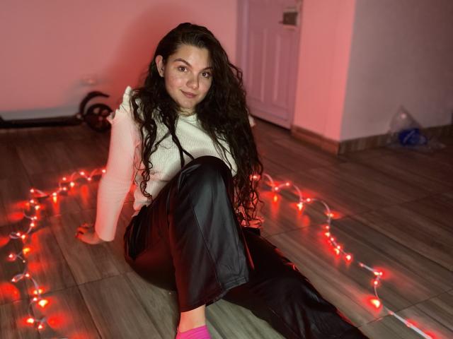 EvaAndMarcelo with long curly black hair, white sleeveless top, black leather pants, pink socks, sitting on wooden floor, surrounded by red Christmas lights, smiling at camera.