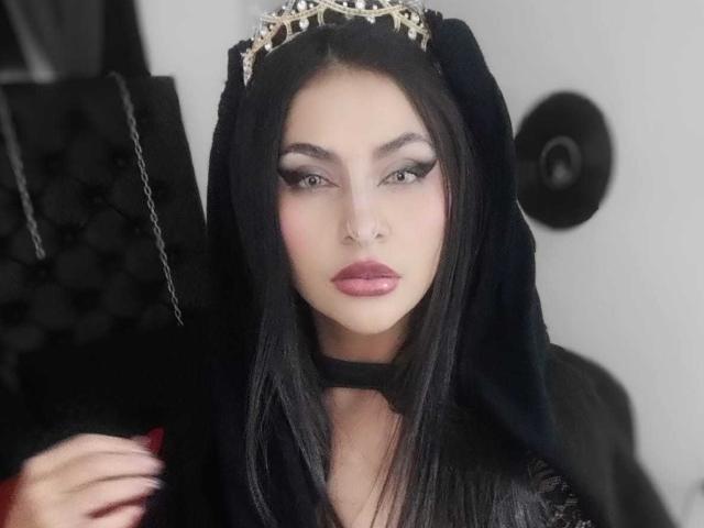 LombanaX, com cabelo preto, usando uma tiara prateada, um capuz preto e um colar, tem maquiagem com olhos esfumados, lábios rosa e uma expressão séria, mostrando unhas pintadas de vermelho..