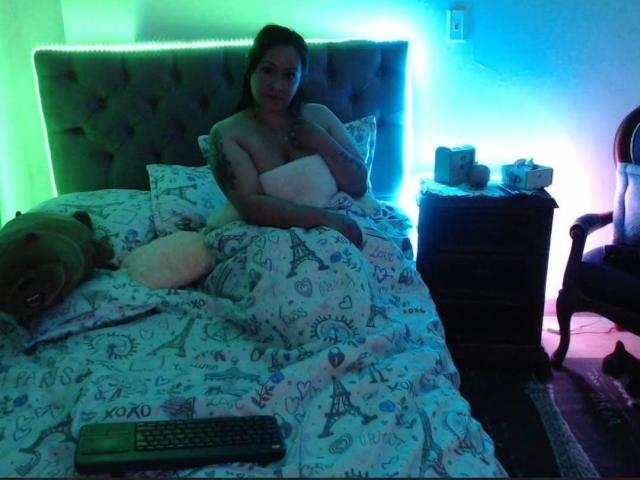 LucianaDia, sitting in a bed with a tufted headboard, has small tits, covered by a white pillow, wearing a white top, with a remote on the bed.