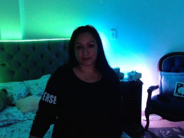 LucianaDia, com cabelo escuro, vestindo um moletom preto com a inscrição "BEST", está em pé em um quarto com pouca luz, com luzes verdes e azuis, e parece um pouco irritada. Quarto com uma cabeceira estofada..