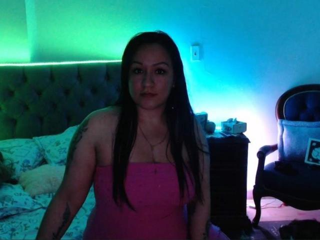 LucianaDia with long black hair, pink strapless dress, and tattoos, sitting in dimly lit bedroom, green and blue light glow, bed with white patterned sheets, wooden chair.
