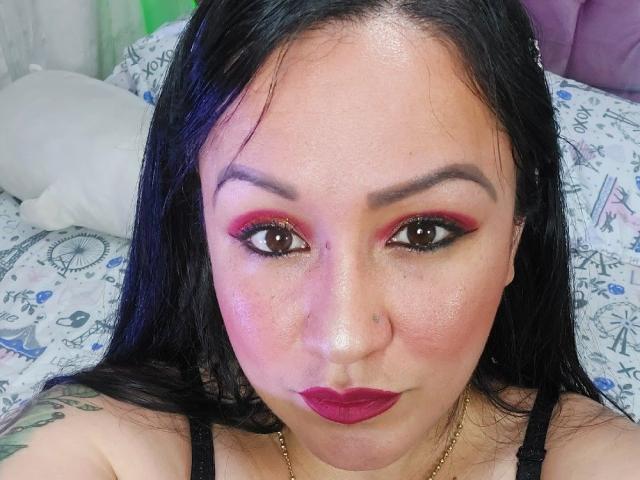 LucianaDia has shiny skin, black hair, red eyeshadow, black eyeliner, pink blush, dark lipstick, and a nose ring. Her tattoos are partially visible on her shoulders.