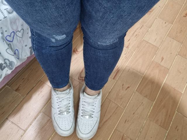 El trasero de Luciana Dia, en unos vaqueros azules ajustados y ligeramente rasgados, está de pie sobre un suelo de madera, llevando puestas zapatillas blancas con cordones, perforadas y de encaje, con los pies muy juntos..