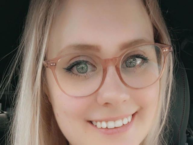 MiraMore-hot tem pele clara, olhos verdes e está sorrindo, com o nariz entre os dentes. Ela está usando óculos com um tom de rosa e tem cabelo loiro. Seus seios não estão visíveis..