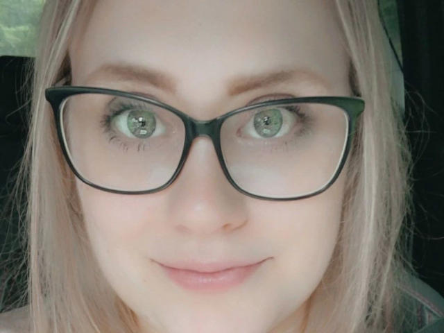 O rosto de MiraMore-hot, com olhos verdes, óculos de aro preto e cabelo loiro. Seus lábios rosados estão ligeiramente curvados em um sorriso discreto. Seus olhos estão levemente cruzados..