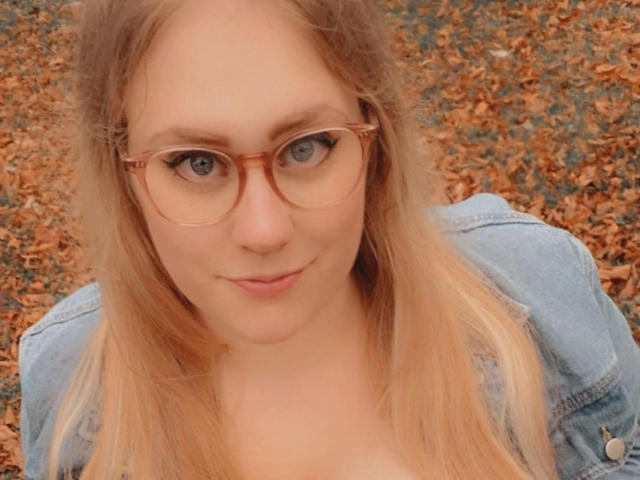 MiraMore-hot with blue glasses, blonde hair, and a denim jacket, showing her tits in a selfie, looking cute with a slight smile.