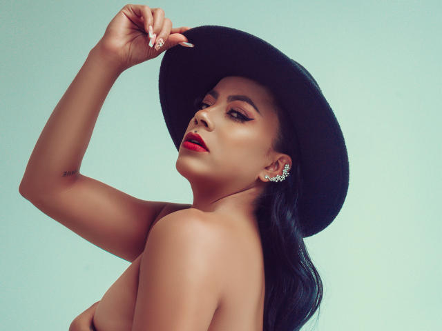 Amberv with dark skin, red lipstick, and long black hair, wearing a black hat, holding it, showing off sparkly earrings, with her bare back and a small tattoo on her arm.