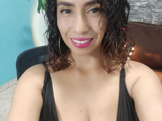 KatyKatt with curly black hair, pink lipstick, and hoop earrings, smiling at the camera, wearing a low-cut black top, showing her boobs. She has a cute, sexy look.