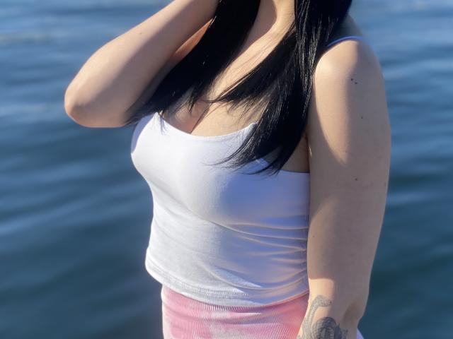 Olieri has black hair, a white tank top showing her small tits, pink pants, and a tattoo on her right arm, standing by blue water.