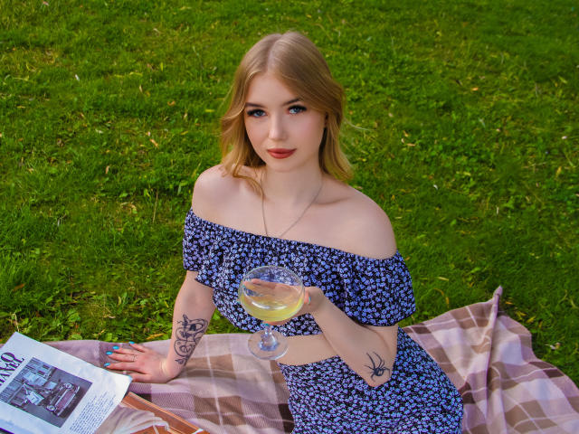 NicoleBrian with blonde hair, blue off-shoulder dress, holding a glass of white wine, sitting on a plaid blanket, showing tattoos on her arm and hand.