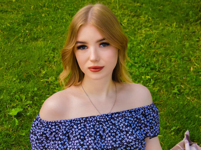 NicoleBrian with blonde hair, blue eyes, and off-shoulder blue floral dress, standing on green grass, wearing a silver necklace, looking directly at camera with a slight smile.