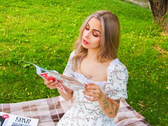 LaraMoss with blonde hair, red lipstick, and floral dress, reading a book on a checkered blanket, showing tattoos on her arm, sitting on green grass.