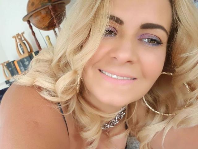 GeilesBabe-hot with blonde curls, purple eyeshadow, and a smile, wearing a silver chain necklace and gold hoop earrings, showing her white teeth and pink lips.