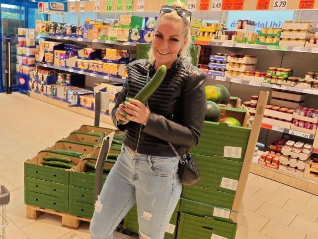 GeilesBabe-hot with blonde hair, black leather jacket, ripped jeans, holding a large green cucumber, smiling in a grocery store, standing in front of green crates with cucumbers.