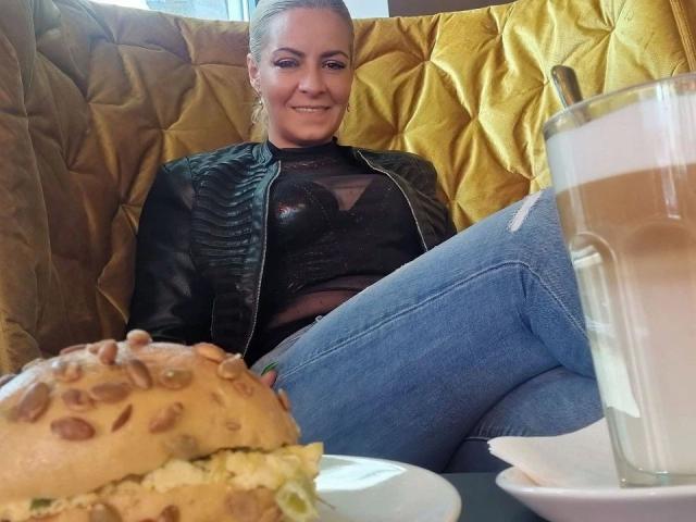 GeilesBabe-hot with blonde ponytail, black leather jacket, and ripped jeans, sitting in a yellow booth, smiling at a large burger and coffee in the foreground.