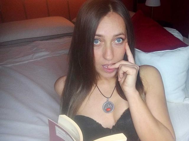 DaisyDea, com olhos azuis, cabelo castanho, sutiã preto e colar, segurando um livro, com o dedo na boca, com uma cama ao fundo, parecendo fofa e sexy, com os seios levemente visíveis..