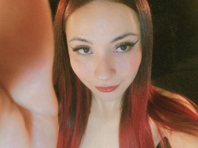 ValeryHarllow with red hair, looking at the camera, has big tits, and is smiling with red lips, thick eyeliner, and a soft, fair skin tone. Her arm is in the foreground.