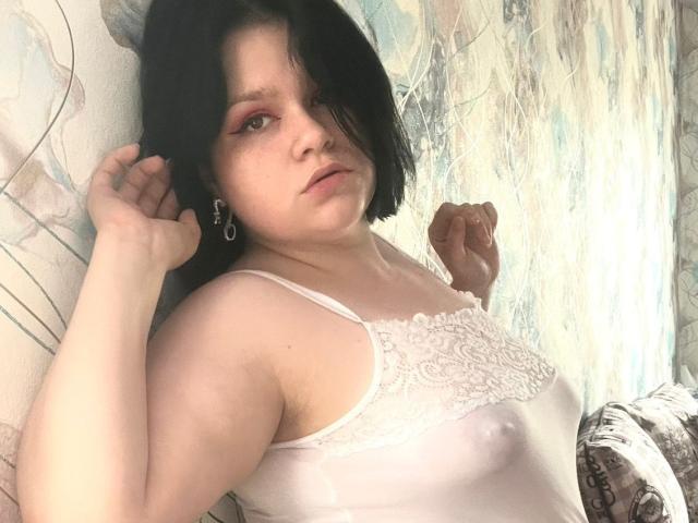 AdelinaOlsen tiene piel pálida, un corte de pelo bob negro, maquillaje rojo en los ojos, una camiseta blanca de encaje, pechos pequeños, pezones visibles, la mano izquierda en el pelo, la mano derecha levantada, mirando a la cámara..