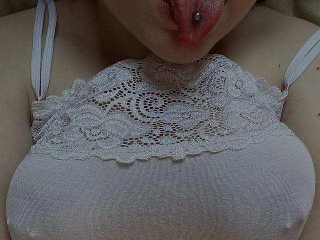 AdelinaOlsen heeft haar tong uitgestoken en draagt een zilveren piercing in haar tong. Ze laat haar bovenborst zien en draagt een witte kanten BH over haar borsten. De bandjes van haar BH zijn zichtbaar op haar schouders..