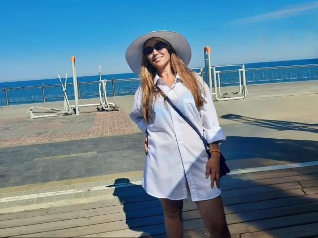 InnaGlory se tient au soleil, vêtue d'une robe blanche, d'un chapeau gris et de lunettes de soleil, avec de longs cheveux blonds, un sac bandoulière noir, et un sourire, sur fond d'un ciel bleu clair..