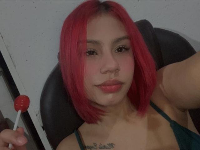 SussyGray with bright pink hair, holding a red lollipop, wearing a green spaghetti strap top, has a tattoo on her chest, sitting in a black chair.